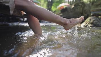 junge Frau, die barfuß im Flusskanal einweicht, Frische aus der Natur, die eine Pause macht, natürliche Ressourcen, Beine schwingt, um das Bachwasser zu spritzen, mit kühlem und frischem Wasser im Bach zu genießen video