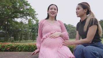 Happy pregnant asian woman and close friend talking laughing while sharing experience of upcoming baby at public green park, two friends having fun with a real life moment, femininity society video