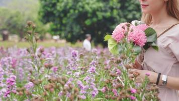 mulher de cabelo loiro de raça muito mista andando dentro de um lindo jardim de flores, segurando flores cor de rosa sentindo-se relaxado e livre ao redor pela natureza, alívio fugir da cidade, sonho feminino ao ar livre video