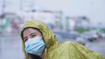 jong mooi Aziatisch meisje draagt beschermend gezichtsmasker, gele regenjas aan de kant van de weg, met auto geparkeerde stadsstraat op de achtergrond, blijf veilig in vervuilde stad, regenseizoen weer klimaat video