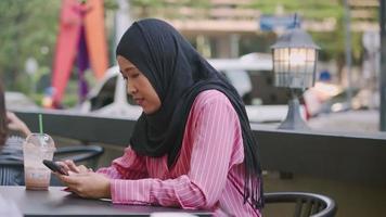 attraente asiatico musulmano seduto da solo sul tavolino da caffè caffè all'aperto utilizzando l'app per cellulare, ottenere messaggi di testo online, social media, leggere sullo schermo del telefono, indossare abiti tradizionali islamici hijab video