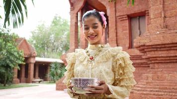 retrata a una mujer hermosa en el festival de songkran con traje tradicional tailandés en el templo sosteniendo un tazón de agua y una sonrisa. cultura de tailandia con festival del agua video