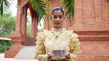 retrata a una mujer hermosa en el festival de songkran con traje tradicional tailandés en el templo sosteniendo un tazón de agua, gira su rostro hacia la cámara y sonríe. cultura de tailandia con festival del agua video