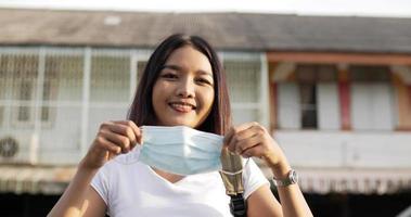 portret van een jonge aziatische vrouw met kort haar die een beschermend medisch gezichtsmasker draagt en op straat staat. veiligheid en gelukkige vrouw die sociale afstand en quarantaine beoefent. zorgconcept. video