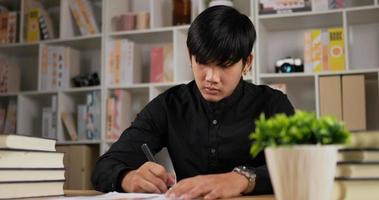 retrato de homem jovem trabalhador asiático focado escrevendo papelada em casa. homem sério trabalhando distante de casa. empresário escrevendo documentos, analisa planos, faz a papelada em um escritório em casa.