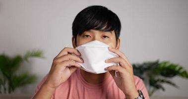 Portrait of Smiling Asian man wearing face mask sits on couch in living room. Happy 20s man putting on protective face masks for covid-19. Coronavirus protection concept.