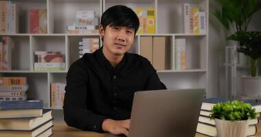 Porträt eines kreativen Asiaten mit Laptop, der im Büro auf die Kamera zeigt. positiver junger geschäftsmann, der kamera betrachtet und lächelt. mann, der an computernotizbuch arbeitet. Geschäfts- und Personenkonzept. video