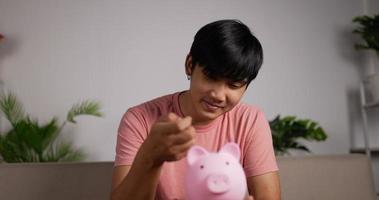 Portrait of Happy man puts coins in piggy bank sits on the couch in living room. Smiling male coins for save home business plan. Saving money is an investment for the future lifestyle. video