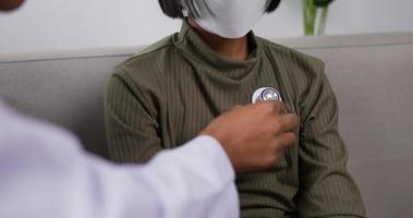 nahaufnahme eines professionellen asiatischen männlichen arztes, der mit einem stethoskop auf das herz eines kleinen mädchens mit gesichtsmaske in der klinik hört. männlicher arzt, der kind untersucht. medizinisches und gesundheitskonzept. video