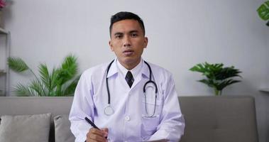 Asian male doctor listening to patient and writing on clipboard while sitting on sofa. Professional physician looking at camera by webcam in web chat consulting client online.Telemedicine concept. video