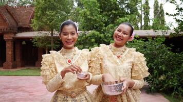 handhållen skott, två vackra kvinnor med thailändsk traditionell dräkt håller vattenskål promenader och leende i templet på songkran festival. thailändska nyåret, thailand kultur med vatten festival video