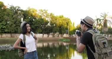 vue latérale d'un couple de voyageurs asiatiques heureux avec un chapeau parlant à la caméra dans le parc. jeune homme prenant des photos pour sa petite amie au parc. concept de passe-temps et de style de vie.