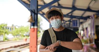 Seitenansicht eines asiatischen jungen Reisenden, der zu Fuß geht und eine Uhr am Bahnhof anschaut. mann mit schutzmasken während des notfalls covid-19. Transport- und Reisekonzept.