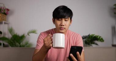 retrato de un joven asiático relajado usando un teléfono inteligente y bebiendo café sentado en el sofá en la sala de estar. navegar en línea leyendo mensajes de redes sociales disfrutando de un estilo de vida cómodo. vacaciones y relax video