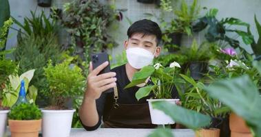 retrato de um jovem jardineiro asiático feliz vendendo on-line nas mídias sociais via smartphone no jardim. homem com máscara facial chamada de vídeo no celular. vegetação em casa, venda on-line e conceito de hobby. video