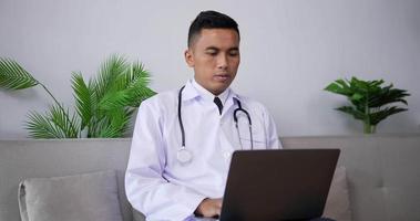 médecin asiatique parlant à un patient faisant un appel vidéo sur un ordinateur portable assis sur un canapé. médecin de sexe masculin de télémédecine parlant en regardant l'ordinateur à écran par webcam dans le client de consultation de chat web en ligne.