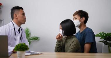 Asian doctor man pediatrician talk to school child girl with face mask make notes in clipboard listen to little patient sit on chair with mother in the hospital. Medical and health care concept.