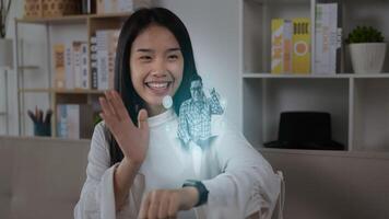 Portrait of Asian woman pressing buttons on smart watch while sitting on sofa. Projecting visible AR screen and chatting on video call. Futuristic and technological concept.