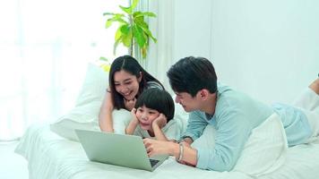 famille heureuse, jeune fils asiatique avec maman et papa allongés sur le lit ensemble. tout en utilisant un ordinateur portable naviguant sur Internet en ligne en regardant des dessins animés, en faisant des achats en ligne, en profitant des médias sociaux pendant les vacances à la maison. video