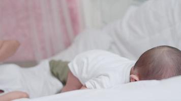 Süßes asiatisches neugeborenes Baby, das auf einem weißen Bett mit einem glücklichen Gesicht spielt. während deine Mutter in der Nähe aufpasst. kleines unschuldiges neugeborenes entzückendes kind am ersten lebenstag. Muttertagskonzept. video