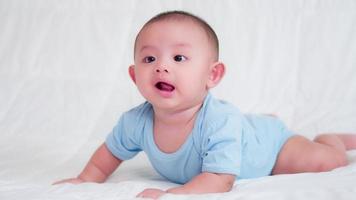 glückliche familie, süßes asiatisches neugeborenes baby tragen blaues hemd liegend, kriechendes spiel auf dem bett, das kamera mit lachendem lächeln glücklichem gesicht betrachtet. unschuldiges kleines neugeborenes entzückendes. elternschaft und muttertagskonzept. video