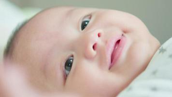 närbild av ansikte söt asiatisk nyfödd baby liggande spela på vit säng titta på kameran med skrattande leende glad ansikte. oskyldigt litet nytt barn bedårande. föräldraskap och mors dag koncept. video