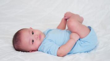 gelukkige familie, schattige aziatische pasgeboren baby draagt blauw shirt liggend spelen op wit bed kijkend naar camera met lachende glimlach blij gezicht. onschuldige kleine nieuwe baby schattig. ouderschap en moederdag concept.