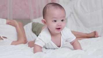 Süßes asiatisches neugeborenes Baby, das auf einem weißen Bett mit einem glücklichen Gesicht spielt. während deine Mutter in der Nähe aufpasst. kleines unschuldiges neugeborenes entzückendes kind am ersten lebenstag. Muttertagskonzept. video