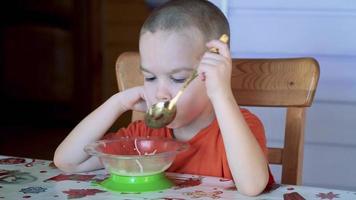 menino de cinco anos sentado na cadeira em uma cozinha e comendo a sopa com a colher. 4k imagens de vídeo em tempo real video