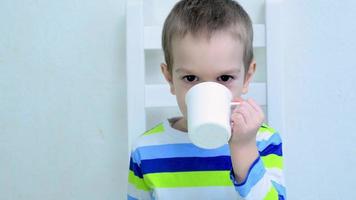 menino de cinco anos sentado em uma cadeira branca e bebendo em uma cozinha. video