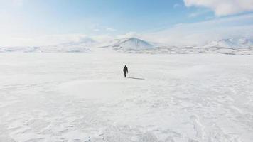 Cinematic aerial view of backpacker walking on frozen lake with beautiful winter mountains scenery background. Solo Adventure lifestyle travel video