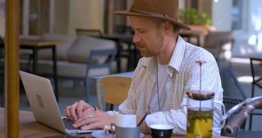 jonge knappe hipster met laptop buiten in het café, bebaarde freelancer werkt in het straatcafé video