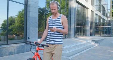 un joven apuesto con una bicicleta colorida y auriculares escucha música y pasea por las calles de verano video