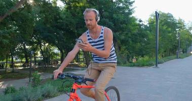 jovem macho bonito com bicicleta colorida e fones de ouvido ouve música e anda pelas ruas de verão video