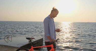 jovem macho bonito em roupas casuais passeio na bicicleta colorida na praia de manhã contra o belo pôr do sol e o mar video