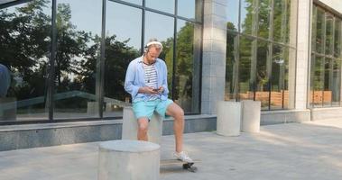 hübscher junger Mann mit Skateboard im Freien in der Stadt Musik mit Kopfhörern hören video