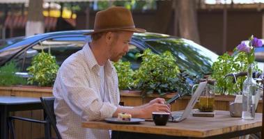 Young handsome hipster with laptop outdoors in the cafe, bearded freelancer work in the street cafe