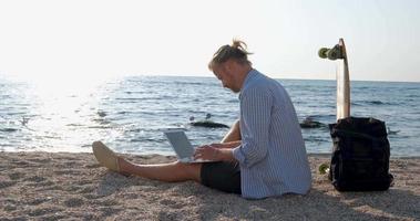 jovem macho bonito na praia de verão trabalha com laptop durante o nascer do sol