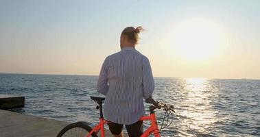 jeune beau mâle en tenue décontractée sur le vélo coloré sur la plage du matin contre le beau coucher de soleil et la mer