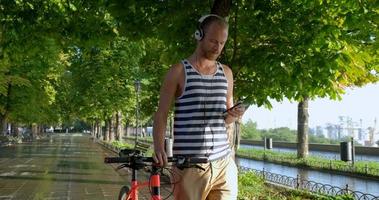 jovem macho bonito com bicicleta colorida e fones de ouvido ouve música e anda pelas ruas de verão video