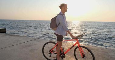 jovem macho bonito em roupas casuais passeio na bicicleta colorida na praia de manhã contra o belo pôr do sol e o mar