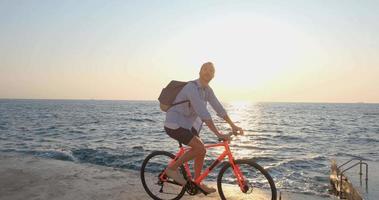 junger gutaussehender mann in freizeitkleidung fährt mit dem bunten fahrrad am morgenstrand gegen den schönen sonnenuntergang und das meer video