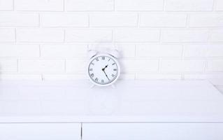 White retro alarm clock with twenty five minutes past one o'clock near brick wall on white workplace. Selective focus photo