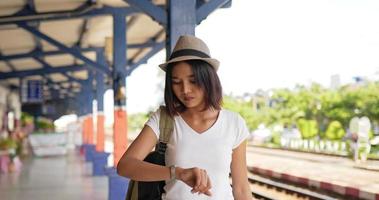 Vista lateral da mulher asiática jovem viajante olhando um relógio e smartphone enquanto caminhava na estação de trem. fêmea usando máscaras protetoras, durante a emergência covid-19. conceito de transporte e viagens. video