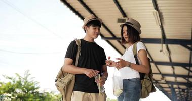 Young Asian traveler couple with hat sausage and drinking water at train station. Happy hungry man and woman eating appetizer. Transportation, travel and food concept.