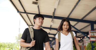 framifrån av lyckliga asiatiska resenärspar med hatt och ryggsäck hand tillsammans medan du går på tågstationen. leende ung man och kvinna ser vy. semester, resor och hobby koncept. video