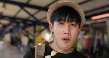 close-up de homem jovem viajante asiático comendo almôndega e olhando para a câmera na estação de trem. feliz macho faminto comendo aperitivo. conceito de transporte, viagens e comida. video