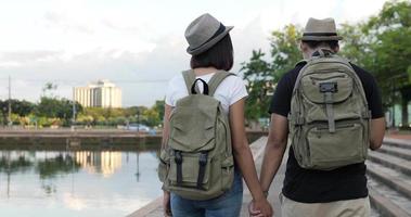 Back view of Happy asian traveler couple with hat and backpack hand together while walking at the park. Smiling young man and woman looking view at park. Holiday, travel and hobby concept.