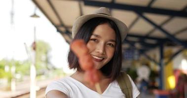 close-up de mulher jovem viajante asiático com chapéu comendo salsicha e olhando para a câmera na estação de trem. feliz fêmea com fome comendo aperitivo. conceito de transporte, viagens e comida. video