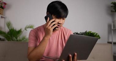 porträt eines asiatischen arbeiters, der einen laptop hält und auf dem smartphone spricht, während er auf einem sofa im wohnzimmer sitzt. Freiberufler, der von zu Hause aus arbeitet. Surfen im Internet, Nutzung sozialer Netzwerke.